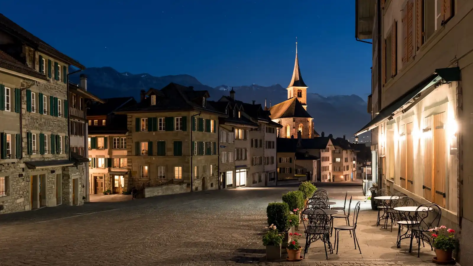 Historische Schweizer Altstadt bei Nacht mit beleuchteten Kirchtürmen, Kopfsteinpflaster und einem ruhigen Strassencafé.