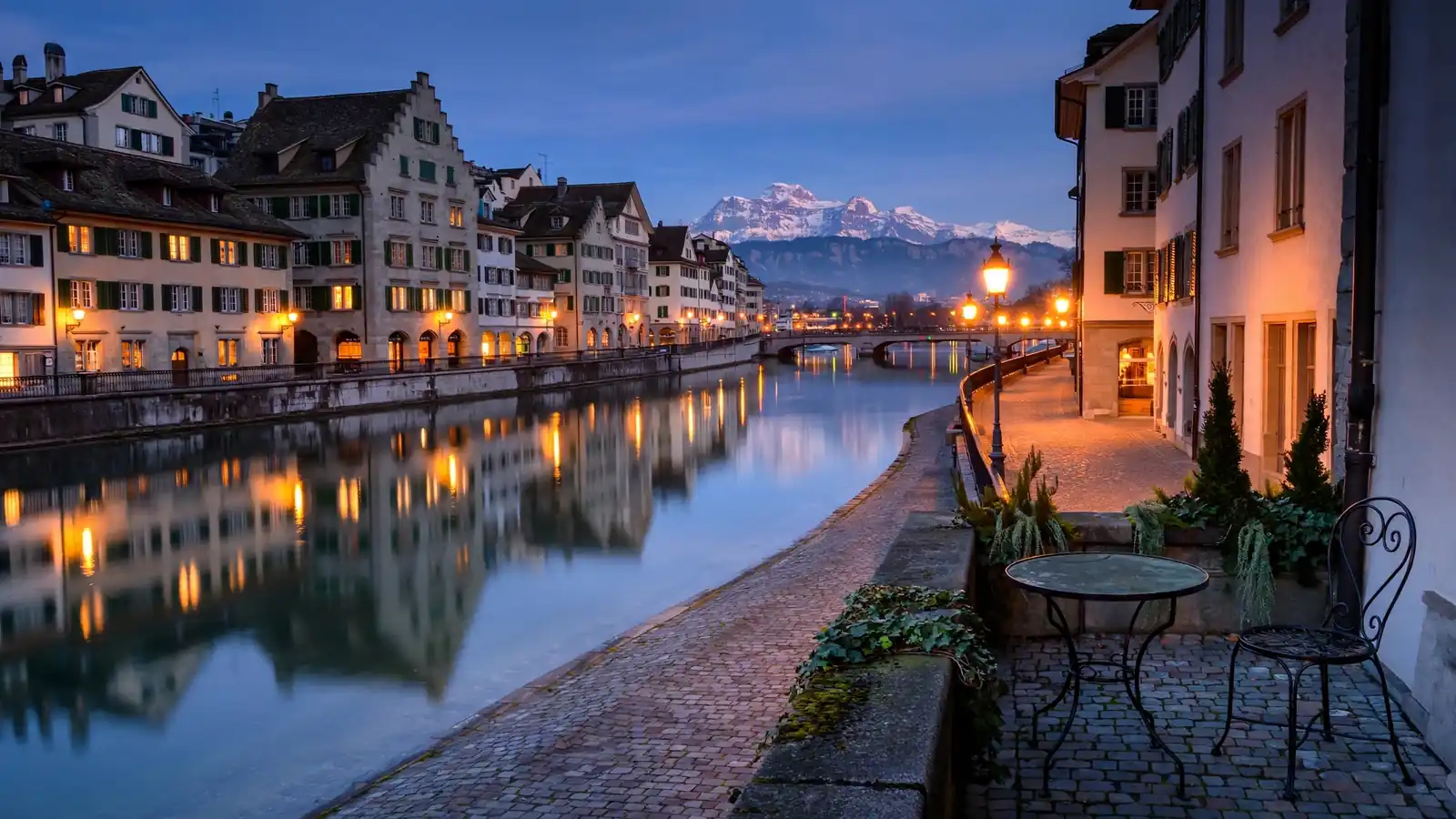Twilight-Blick auf die Zürcher Altstadt entlang der Limmat mit Alpen und einem leeren Bistro-Stuhl.